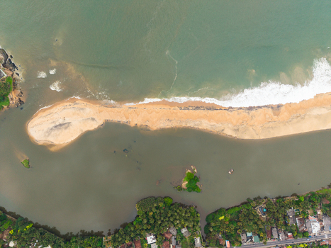 Aerial view of a narrow sandbar separating the turquoise ocean from a calm river mouth lined with tropical trees and buildings in Galle Southern Province Sri Lanka. Aerial view of a narrow sandbar separating the turquoise ocean from a calm river mouth lined with tropical trees and buildings in Galle Southern Province Sri Lanka.