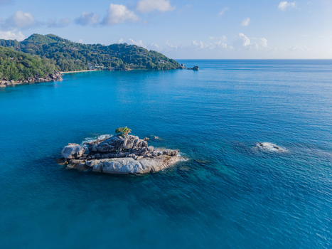 Aerial view of a small rocky island topped with sparse greenery surrounded by the deep blue of the Indian Ocean near lush green hills Victoria Seychelles. Aerial view of a small rocky island topped with sparse greenery surrounded by the deep blue of the Indian Ocean near lush green hills Victoria Seychelles.