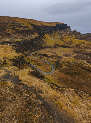 Aerial view of The Quiraing featuring a winding road through steep rocky cliffs and golden brown moorland under a cloudy sky Isle of Skye Scotland United Kingdom. Aerial view of The Quiraing featuring a winding road through steep rocky cliffs and golden brown moorland under a cloudy sky Isle of Skye Scotland United Kingdom.