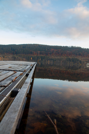 View of a wooden pier on a calm lake reflecting the autumn colors of the surrounding forested hills under a soft blue sky Breitnau Baden Wurttemberg Germany. View of a wooden pier on a calm lake reflecting the autumn colors of the surrounding forested hills under a soft blue sky Breitnau Baden Wurttemberg Germany.