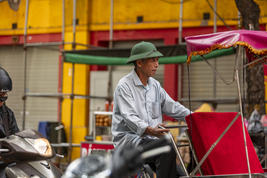 Hanoi Ha Noi Vietnam 11 November 2025 View of a man in a pith helmet driving a cyclo through a busy street with yellow buildings and scaffolding in the background.