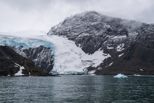 View of a glacier flowing down a rocky snow dusted mountain into the sea with a small iceberg floating in the water in the South Orkney Islands. View of a glacier flowing down a rocky snow dusted mountain into the sea with a small iceberg floating in the water in the South Orkney Islands.