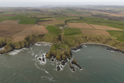 Aerial view of Dunnottar Castle a ruined medieval fortress on a rocky cliff edge overlooking the North Sea with surrounding green fields in Stonehaven Scotland United Kingdom. Aerial view of Dunnottar Castle a ruined medieval fortress on a rocky cliff edge overlooking the North Sea with surrounding green fields in Stonehaven Scotland United Kingdom.
