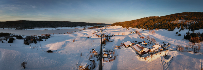 Aerial view of a serene snow blanketed landscape dotted with rustic buildings and a distant lake under a soft golden light Les Rousses Bourgogne Franche Comté France. Aerial view of a serene snow blanketed landscape dotted with rustic buildings and a distant lake under a soft golden light Les Rousses Bourgogne Franche Comté France.