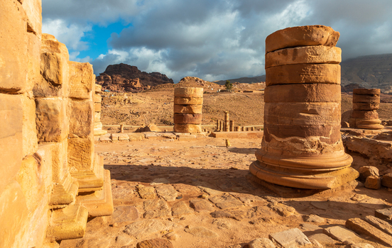View of ancient stone columns and ruins of the Great Temple in the desert landscape of Petra with rocky mountains under a cloudy sky in Wadi Musa Ma an Governorate Jordan. View of ancient stone columns and ruins of the Great Temple in the desert landscape of Petra with rocky mountains under a cloudy sky in Wadi Musa Ma an Governorate Jordan.