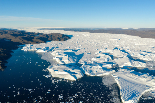 Aerial view of Ilulissat glacier with massive icebergs floating in dark blue water surrounded by rugged rocky coastlines under a clear blue sky in Greenland. Aerial view of Ilulissat glacier with massive icebergs floating in dark blue water surrounded by rugged rocky coastlines under a clear blue sky in Greenland.