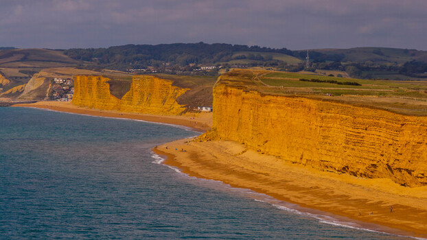 Aerial view of West Bay featuring iconic golden sandstone cliffs along the Jurassic Coast with sandy beach and rolling green hills in West Bay England United Kingdom. Aerial view of West Bay featuring iconic golden sandstone cliffs along the Jurassic Coast with sandy beach and rolling green hills in West Bay England United Kingdom.