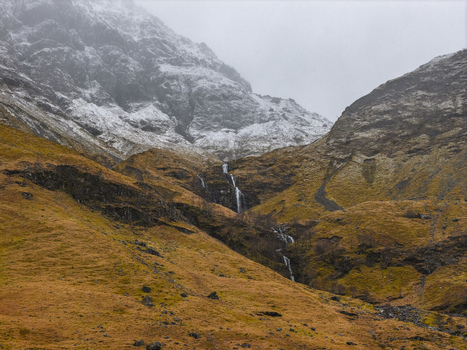 Aerial view of the rugged Glencoe valley with a cascading waterfall snow dusted mountain peaks and dramatic orange slopes under a misty sky in Glencoe Scotland United Kingdom. Aerial view of the rugged Glencoe valley with a cascading waterfall snow dusted mountain peaks and dramatic orange slopes under a misty sky in Glencoe Scotland United Kingdom.