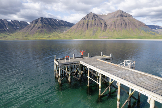 Aerial view of Onundarfjordur Pier with a person in an orange jacket standing on the wooden structure over calm water against a backdrop of mountains in Holt Isafjardabaer Iceland. Aerial view of Onundarfjordur Pier with a person in an orange jacket standing on the wooden structure over calm water against a backdrop of mountains in Holt Isafjardabaer Iceland.