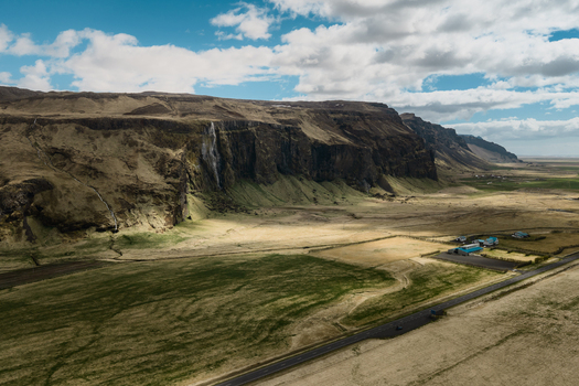 Aerial view of Seljalandsfoss waterfall cascading down dramatic cliffs surrounded by vast green fields a winding road and scattered buildings in Rangarthing eystra Iceland. Aerial view of Seljalandsfoss waterfall cascading down dramatic cliffs surrounded by vast green fields a winding road and scattered buildings in Rangarthing eystra Iceland.
