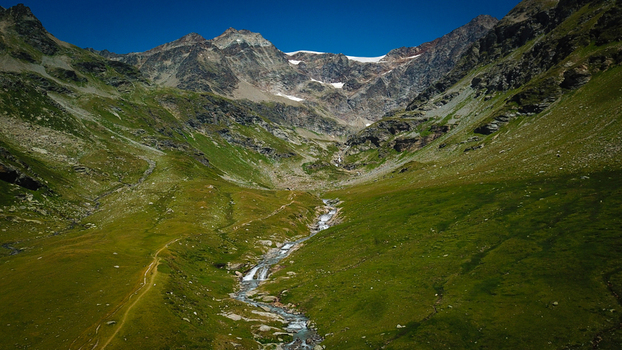Aerial view of a meandering stream cutting through verdant meadows towards rugged snow capped peaks Lago di San Grato Valgrisenche Aosta Valley Italy.