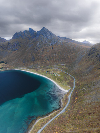 Aerial view of the dramatic mountains and turquoise waters of a fjord in Senja with a coastal road winding along the shoreline under a cloudy sky in Senja Troms Norway.
