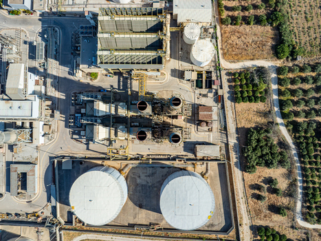 Aerial view of a power plant facility featuring large storage tanks industrial cooling towers and complex piping systems situated near olive groves in Chania Greece.