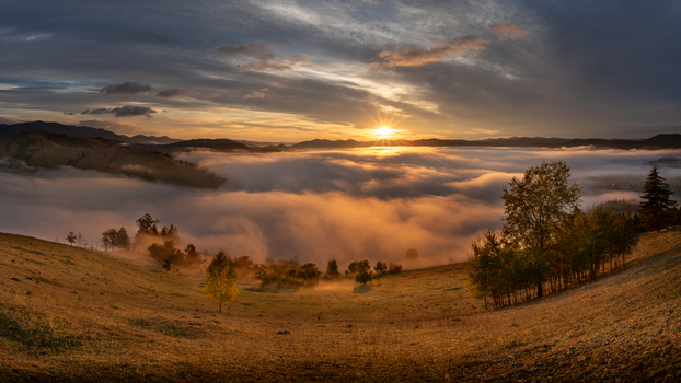 View of the sunrise over a misty valley in the Carpathian Mountains with golden light illuminating the rolling hills and autumn trees in Moldovita Suceava County Romania. View of the sunrise over a misty valley in the Carpathian Mountains with golden light illuminating the rolling hills and autumn trees in Moldovita Suceava County Romania.