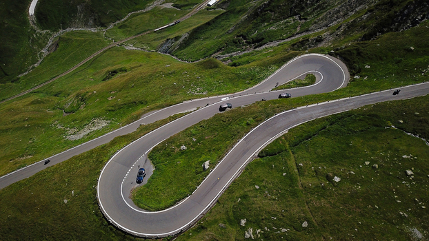 Aerial view of the winding hairpin turns of the Furka Pass mountain road with cars driving through lush green alpine slopes and a railway line in Obergoms Valais Switzerland. Aerial view of the winding hairpin turns of the Furka Pass mountain road with cars driving through lush green alpine slopes and a railway line in Obergoms Valais Switzerland.