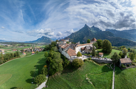 Aerial view of the historic Chateau de Gruyeres perched on a hill surrounded by lush green meadows traditional architecture and Alpine peaks in Gruyeres Fribourg Switzerland. Aerial view of the historic Chateau de Gruyeres perched on a hill surrounded by lush green meadows traditional architecture and Alpine peaks in Gruyeres Fribourg Switzerland.