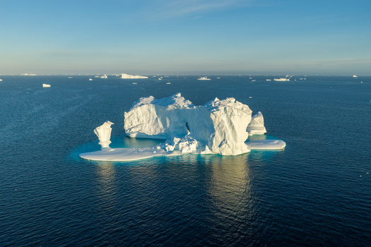 Aerial view of a massive arch iceberg floating in the deep blue waters of the Arctic Ocean surrounded by smaller ice fragments under a clear sky in Greenland. Aerial view of a massive arch iceberg floating in the deep blue waters of the Arctic Ocean surrounded by smaller ice fragments under a clear sky in Greenland.
