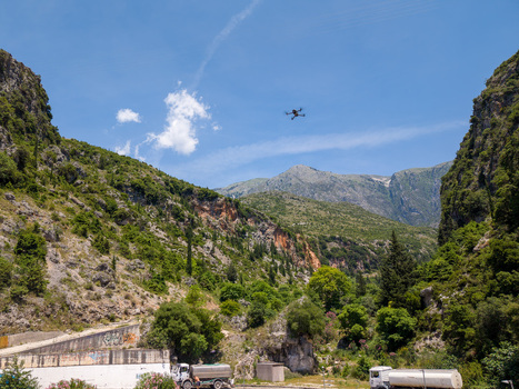 Aerial view of a drone flying through a mountain valley with rocky cliffs green vegetation and industrial trucks under a clear blue sky in Dhermi Vlore County Albania.