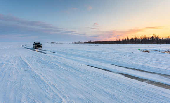View of a car driving on the frozen surface of Lake Baikal with tire tracks in the snow distant trees and a soft sunset sky in Severobaykalsk Buryatia Russia. View of a car driving on the frozen surface of Lake Baikal with tire tracks in the snow distant trees and a soft sunset sky in Severobaykalsk Buryatia Russia.