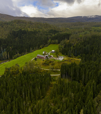 Aerial view of a traditional farmstead with red and white houses nestled in a lush green clearing surrounded by dense evergreen forests and rolling hills in Skare Vestland Norway.