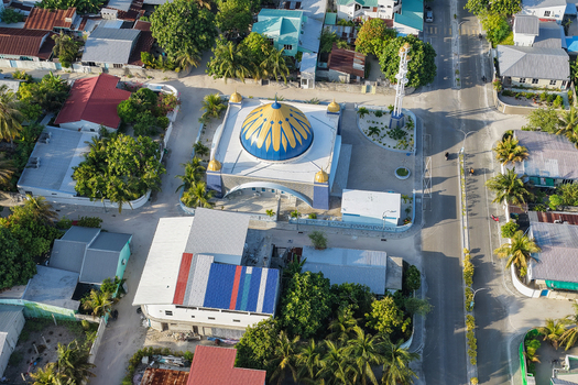 Aerial view of a mosque with a distinctive blue and gold dome surrounded by residential buildings lush tropical trees and paved streets in Kudahuvadhoo Dhaalu Atoll Maldives.