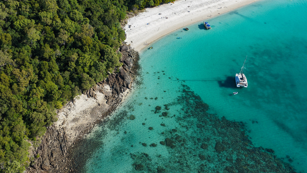 Aerial view of Whitehaven Beach with turquoise water coral reefs a catamaran anchored in the bay and lush tropical forest along the rocky coastline in Whitsundays Queensland Australia.