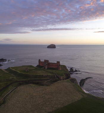 Aerial view of Tantallon Castle ruins on a cliff overlooking the sea with Bass Rock in the distance under a sunset sky North Berwick Scotland United Kingdom.