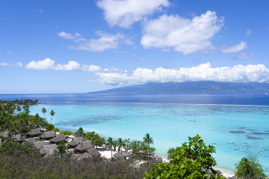 View of tropical bungalows nestled along a white sand beach with turquoise lagoon waters and lush mountains in the background in Moorea Maiao Windward Islands French Polynesia.