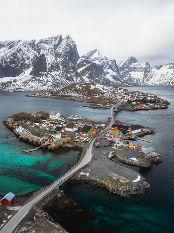 Aerial view of quaint yellow houses nestled on rocky islands connected by bridges over turquoise waters with snow dusted mountains looming in the distance Sakrisøya Nordland Norway. Aerial view of quaint yellow houses nestled on rocky islands connected by bridges over turquoise waters with snow dusted mountains looming in the distance Sakrisøya Nordland Norway.