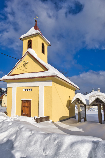 View of a small vibrant yellow chapel with a red roof stands nestled in the snow against a bright blue sky Polianka Banskobystricky kraj Slovakia. View of a small vibrant yellow chapel with a red roof stands nestled in the snow against a bright blue sky Polianka Banskobystricky kraj Slovakia.