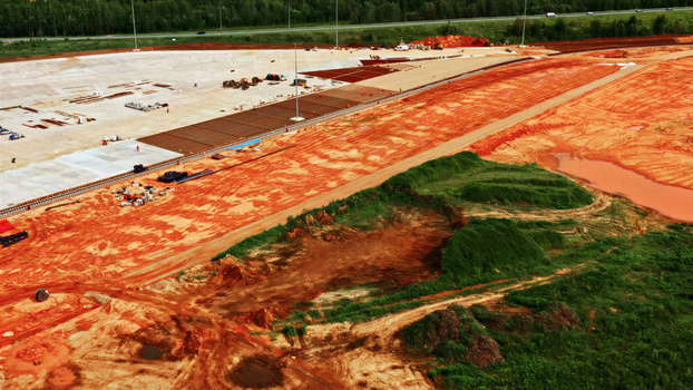 Aerial view of a large construction site featuring exposed red soil concrete foundations and heavy machinery with a highway visible in the background in Latvia. Aerial view of a large construction site featuring exposed red soil concrete foundations and heavy machinery with a highway visible in the background in Latvia.