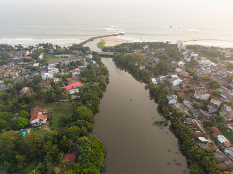 Aerial view of a wide river flowing through a coastal town with lush tropical greenery and residential houses towards the ocean in Galle Southern Province Sri Lanka.