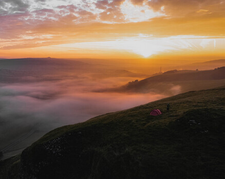 Aerial view of Winnats Pass at sunrise with a small tent and a person on a grassy hillside overlooking a valley filled with morning mist in Hope Valley England United Kingdom. Aerial view of Winnats Pass at sunrise with a small tent and a person on a grassy hillside overlooking a valley filled with morning mist in Hope Valley England United Kingdom.