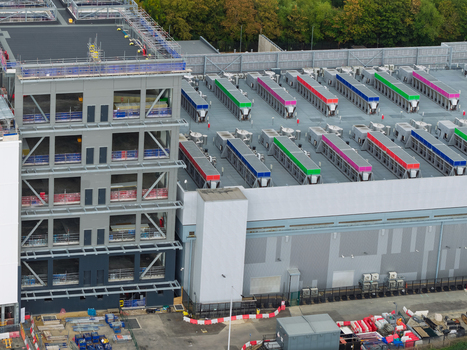 Aerial view of a modern data center featuring a multi story steel framed building and rows of colorful industrial cooling units on the roof in the United Kingdom.