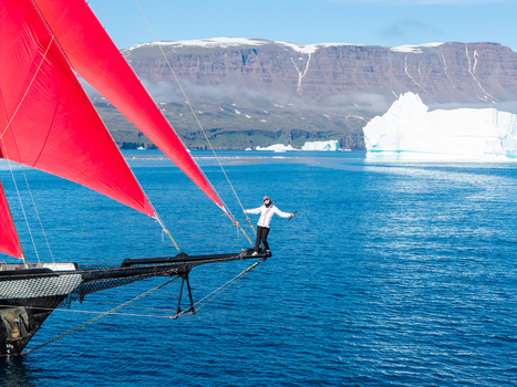Greenland 30 June 2026 Aerial view of a person standing on the bowsprit of a sailing ship with vibrant red sails surrounded by blue arctic waters a large iceberg and distant snow capped mountains. Greenland 30 June 2026 Aerial view of a person standing on the bowsprit of a sailing ship with vibrant red sails surrounded by blue arctic waters a large iceberg and distant snow capped mountains.