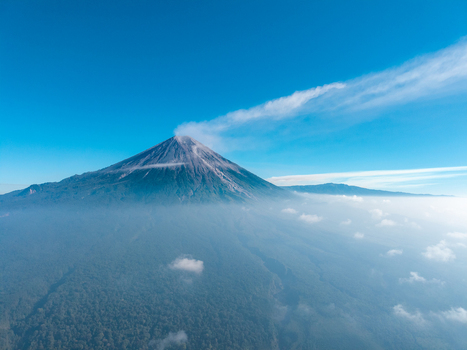 Aerial view of Mount Semeru with a volcanic smoke plume rising above lush forested slopes and a sea of clouds under a clear blue sky in Pasrujambe East Java Indonesia. Aerial view of Mount Semeru with a volcanic smoke plume rising above lush forested slopes and a sea of clouds under a clear blue sky in Pasrujambe East Java Indonesia.