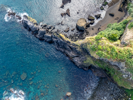 Aerial view of rugged volcanic cliffs and a black sand beach meeting turquoise ocean waters in Ribeira Grande Azores Portugal.