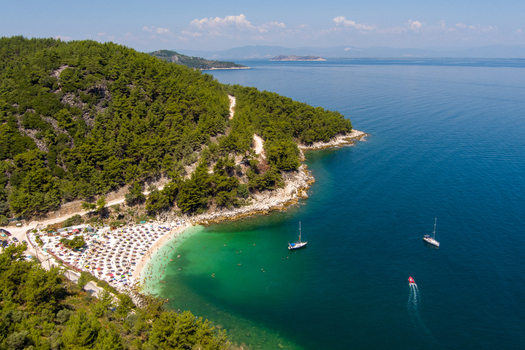 Greece 21 September 2026 Aerial view of a secluded beach with turquoise waters sun umbrellas and pine covered hills along the coastline with sailboats in the sea. Greece 21 September 2026 Aerial view of a secluded beach with turquoise waters sun umbrellas and pine covered hills along the coastline with sailboats in the sea.