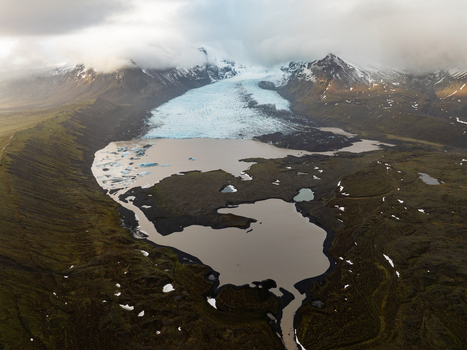 Aerial view of Kviarjokull glacier tongue flowing into a glacial lagoon with dark volcanic sediment and surrounding mossy mountains in Sveitarfelagid Hornafjordur Iceland.