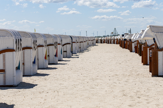 View of rows of traditional white wicker hooded beach chairs on the sandy Strand Schillig under a bright sky with wind turbines in the distance Wangerland Niedersachsen Germany. View of rows of traditional white wicker hooded beach chairs on the sandy Strand Schillig under a bright sky with wind turbines in the distance Wangerland Niedersachsen Germany.