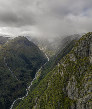 Aerial view of the Buarbreen glacier and a deep mountain valley with a winding river and steep rocky cliffs under a misty sky in Ullensvang Vestland Norway. Aerial view of the Buarbreen glacier and a deep mountain valley with a winding river and steep rocky cliffs under a misty sky in Ullensvang Vestland Norway.