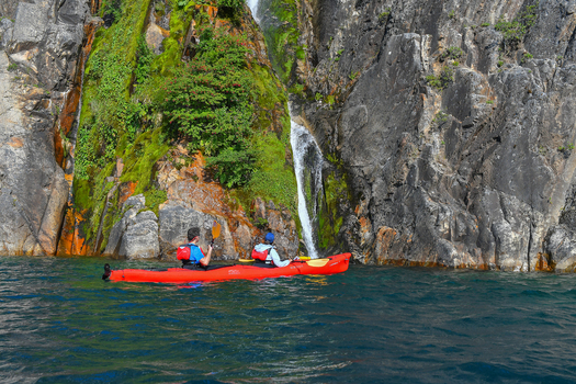 View of two people in a red kayak paddling on the blue waters of Nahuel Huapi Lake near a small waterfall and rocky cliffs San Carlos de Bariloche Rio Negro Argentina. View of two people in a red kayak paddling on the blue waters of Nahuel Huapi Lake near a small waterfall and rocky cliffs San Carlos de Bariloche Rio Negro Argentina.