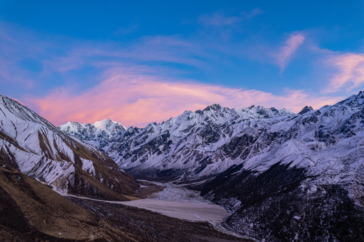 Aerial view of snow capped Himalayan mountain peaks under a vibrant pink and blue sunset sky with a rugged valley floor below in Nepal.