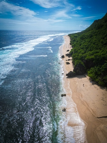 Aerial view of Pantai Nunggalan with its sandy beach rocky cliffs lush greenery and turquoise ocean waves in Kecamatan Kuta Selatan Bali Indonesia.