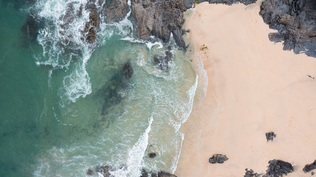 Aerial view of Harlyn Bay Beach featuring turquoise waves crashing against dark jagged rocks and golden sand Saint Merryn Cornwall United Kingdom.