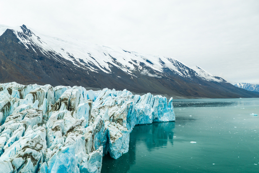 Aerial view of a massive glacier calving into the turquoise sea with snow capped mountains in the background in Greenland.