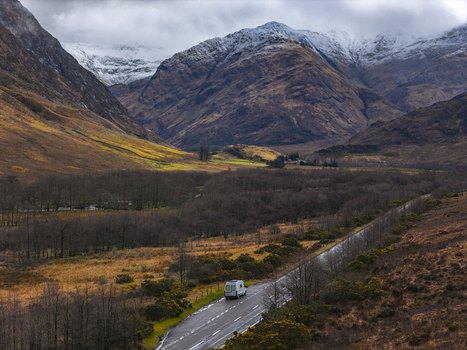 Aerial view of a white camper van driving on a winding road through the rugged Glencoe valley with snow capped mountains and dramatic clouds in Scotland United Kingdom. Aerial view of a white camper van driving on a winding road through the rugged Glencoe valley with snow capped mountains and dramatic clouds in Scotland United Kingdom.
