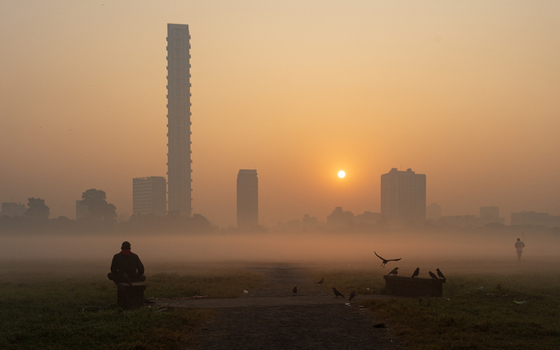 View of a man sitting on a bench in a misty park during sunrise with the Kolkata skyline and The 42 skyscraper in the background in Kolkata West Bengal India. View of a man sitting on a bench in a misty park during sunrise with the Kolkata skyline and The 42 skyscraper in the background in Kolkata West Bengal India.