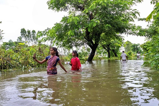 Banshkhali Upazila Chittagong Division Bangladesh 10 July 2026 View of two young girls wading through floodwaters in a rural area with lush trees and a man walking in the background. Banshkhali Upazila Chittagong Division Bangladesh 10 July 2026 View of two young girls wading through floodwaters in a rural area with lush trees and a man walking in the background.