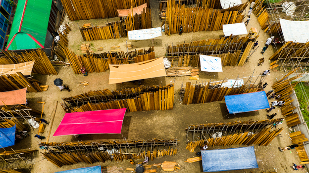 Aerial view of Baish Mowza Sarak Bazar with rows of stacked timber planks and colorful tarps people walking through the market in Nabinagar Chittagong Division Bangladesh. Aerial view of Baish Mowza Sarak Bazar with rows of stacked timber planks and colorful tarps people walking through the market in Nabinagar Chittagong Division Bangladesh.